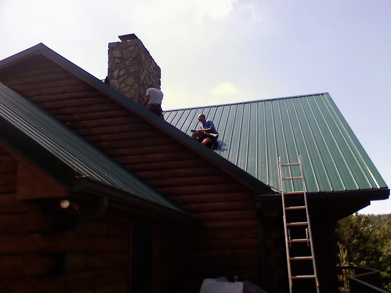 Roofers installing green metal panels beside a stone chimney on a log home in Farmington